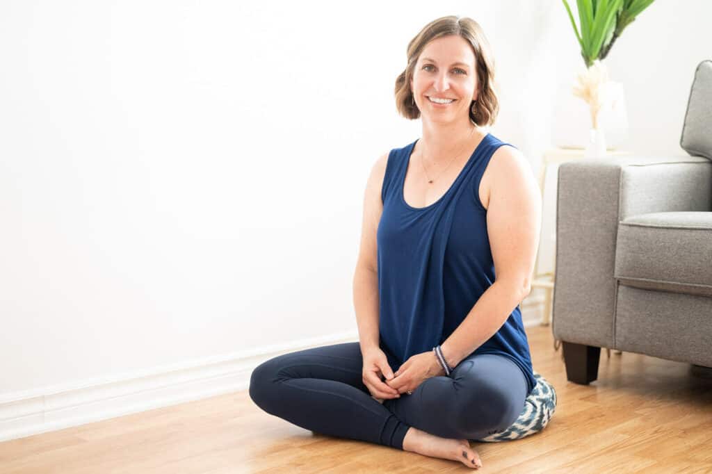 Beth Hoag, a cancer rehabilitation physiotherapist, is sitting on the ground with her legs crossed, smiling and wearing a blue tank top and leggings.
