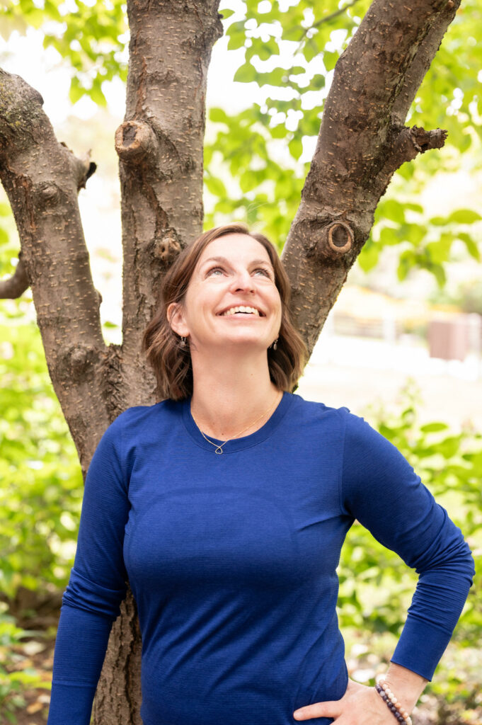Beth Hoag, cancer rehab physiotherapist in Ottawa, ON, is standing under a tree looking up wearing a blue shirt and smiling.
