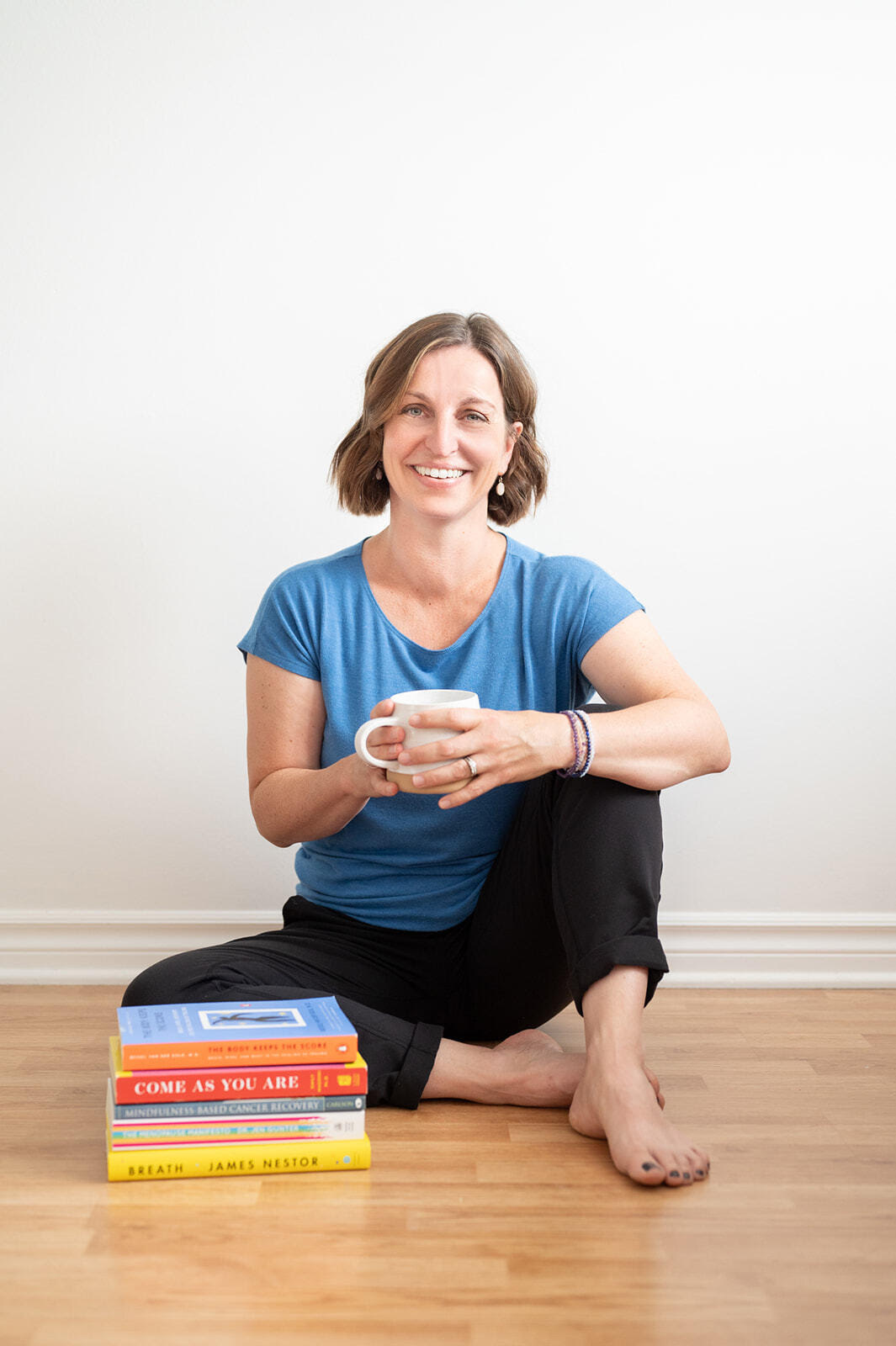 Beth Hoag, cancer rehab physiotherapist is sitting on the floor with a stack of books in front of her smiling and holding a coffee cup.