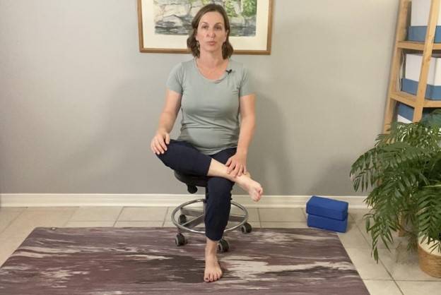 Beth Hoag, Physiotherapist, sitting on a stool with one leg crossed over her opposite thing in a figure 4 position, a beneficial hip and low back stretch after pelvic radiation therapy.