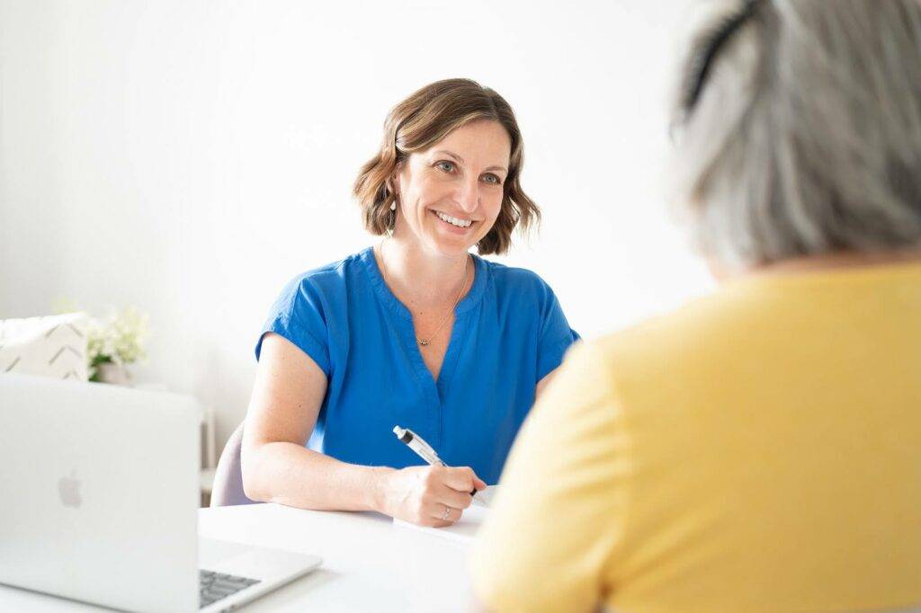 Beth Hoag, PT, CLT Registered Physiotherapist, is sitting across from a patient discussing solutions for hip and low back pain after pelvic radiation treatment.